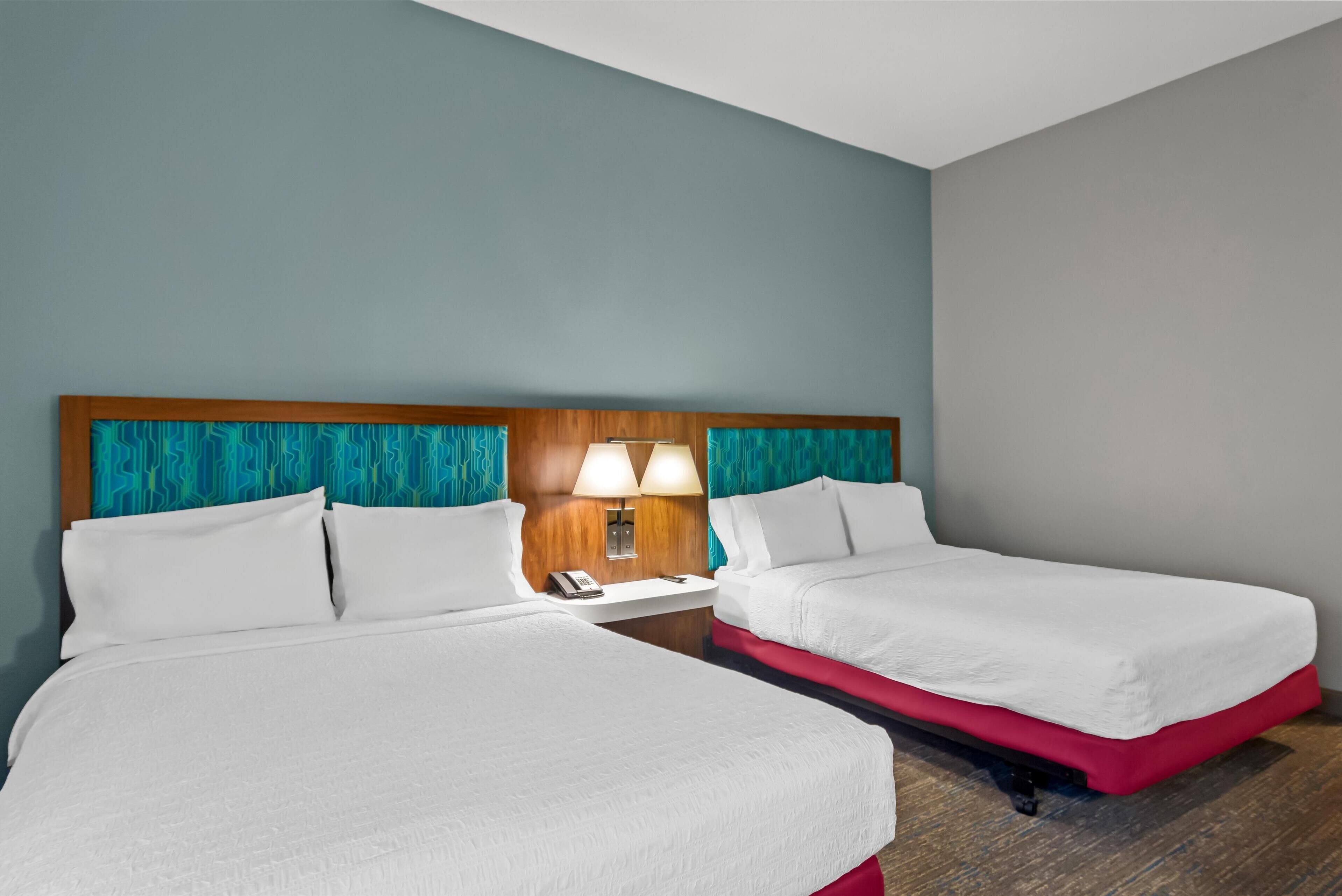A hotel room with two beds, a wooden headboard, teal curtains, a nightstand with a lamp, and a phone.