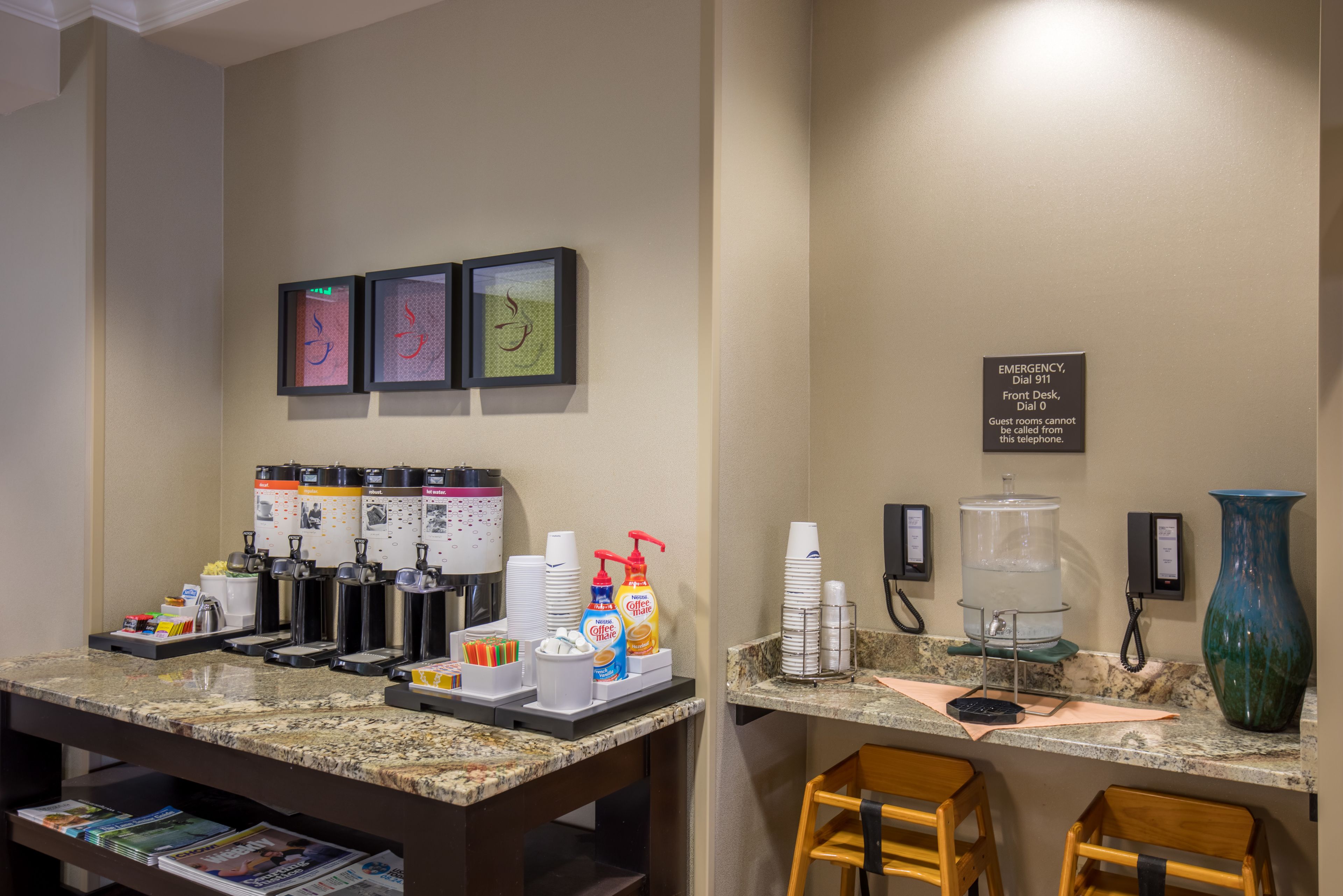 A hotel reception area with a coffee station, framed artwork, and a sign indicating the emergency phone number.