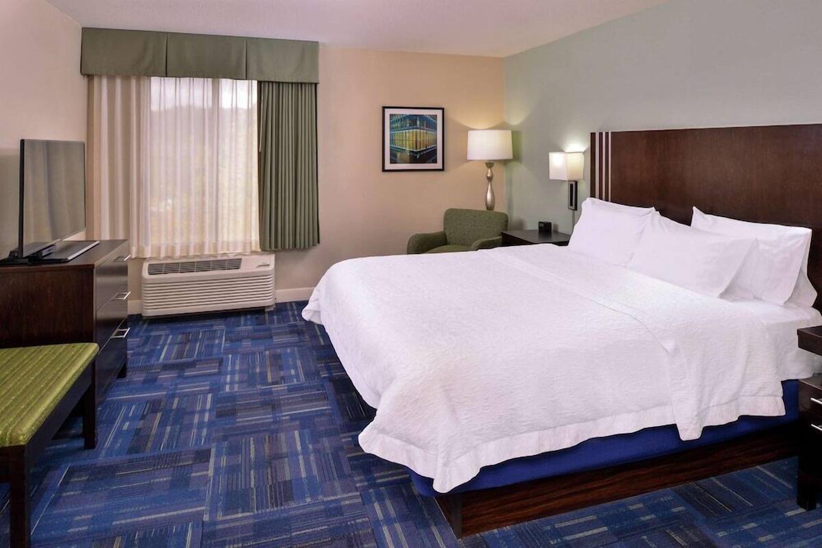 A hotel room with a large bed, a desk with a chair, a standing heater, a bench, and a window with curtains.