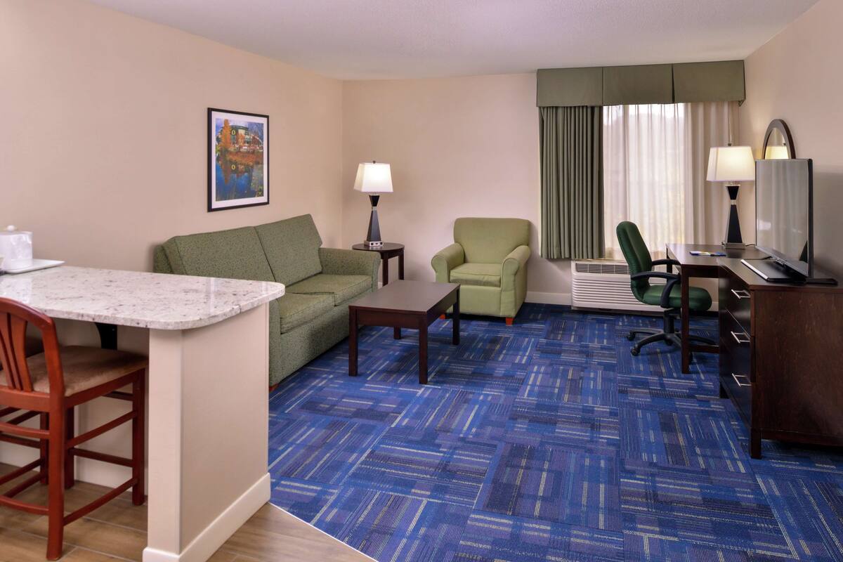 A hotel room with a living area, a dining table, a sofa, a chair, a desk with a computer, and a TV.