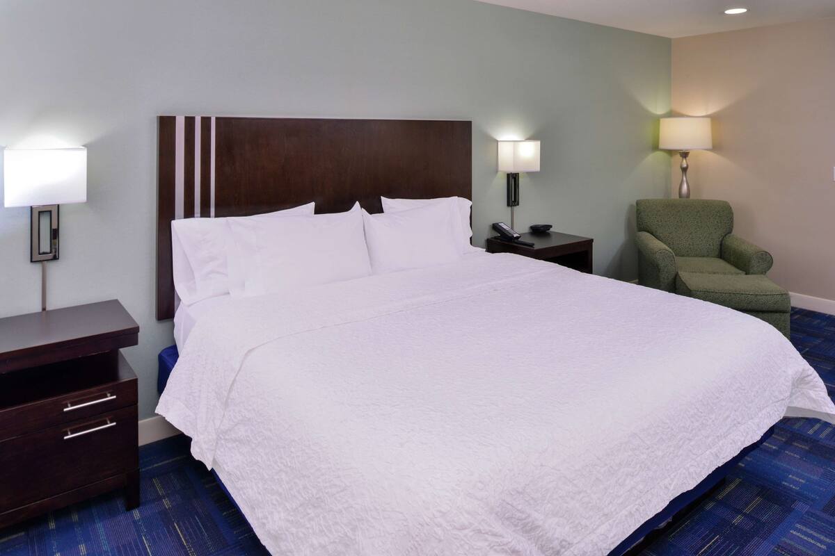 A hotel room with a large bed, a wooden headboard, a green armchair, a nightstand with a lamp, and a desk with drawers.