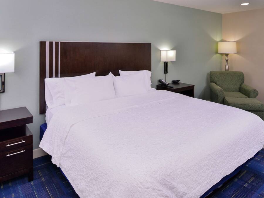 Een hotelkamer met een groot bed, een houten hoofdbord, een groene fauteuil, een nachtkastje met lamp en een bureau met lades.