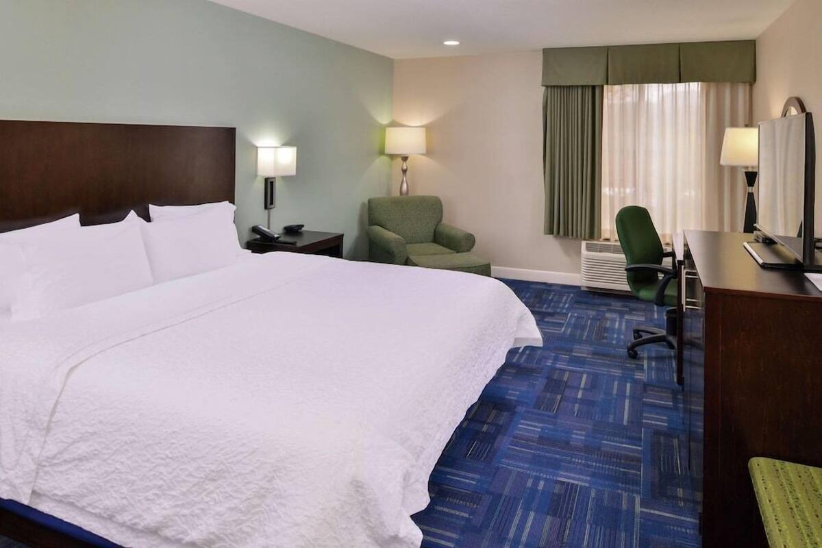 A hotel room with a large bed, a desk with a computer, a chair, a lamp, and a window with curtains.