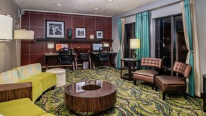A hotel room with a green patterned carpet, a round wooden coffee table, a sofa, a chair, and a desk with a computer. - Hampton Inn Eugene (Eugene)