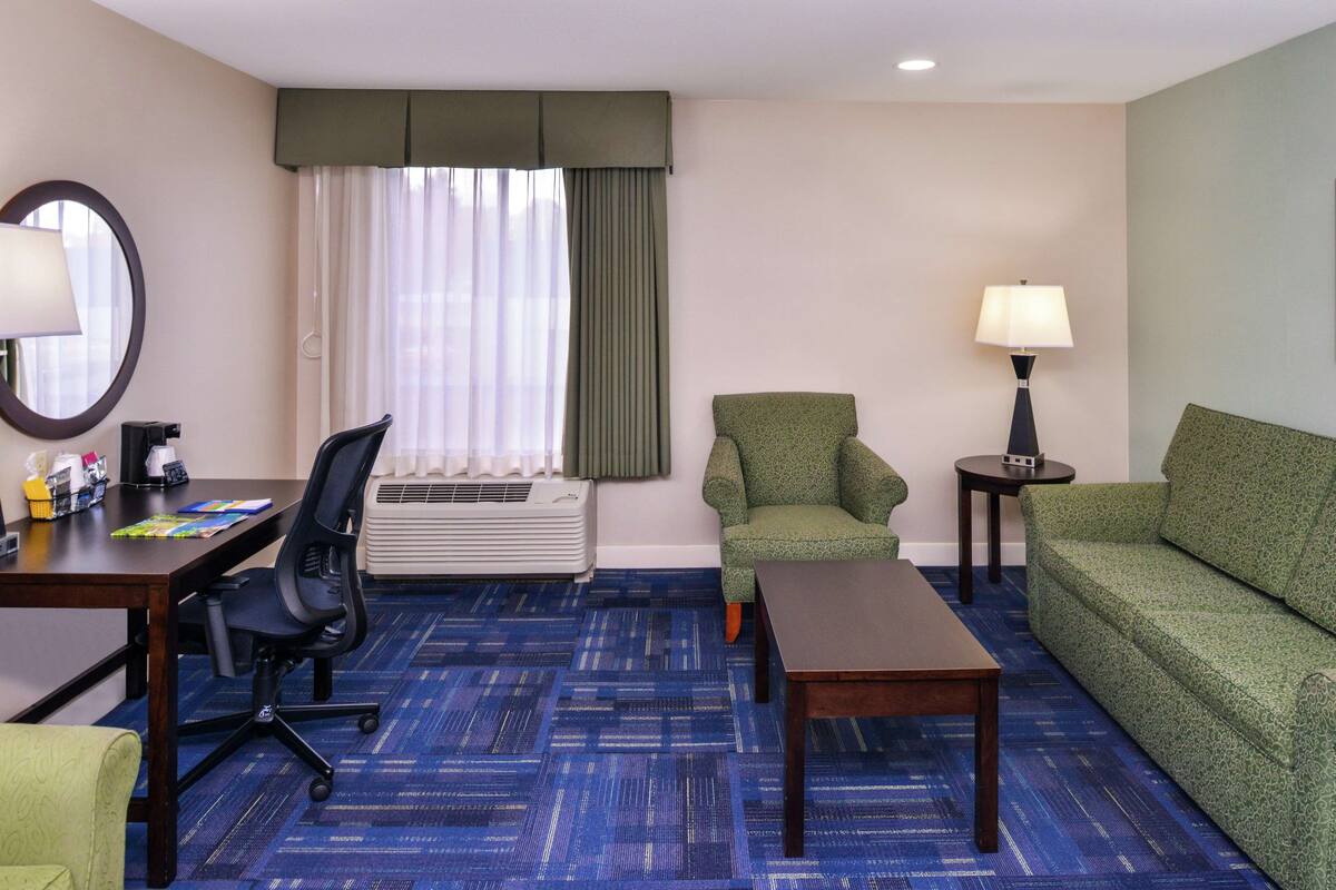 A hotel room with a blue carpet, a green sofa, a green armchair, a wooden coffee table, a desk with a chair, a lamp, a window with curtains, and an air conditioning unit.