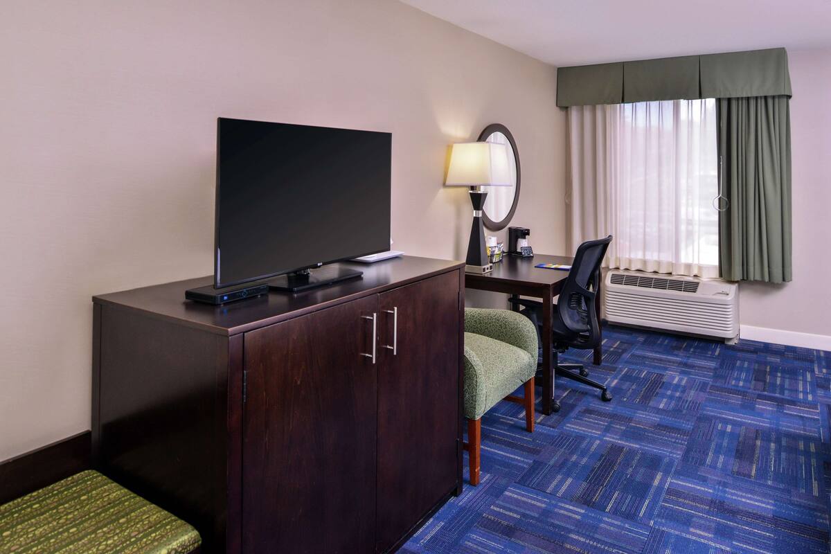 A hotel room with a flat-screen TV, a desk with a chair, a lamp, and a large window with curtains.