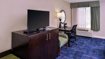 A hotel room with a flat-screen TV, a desk with a chair, a lamp, and a large window with curtains.
