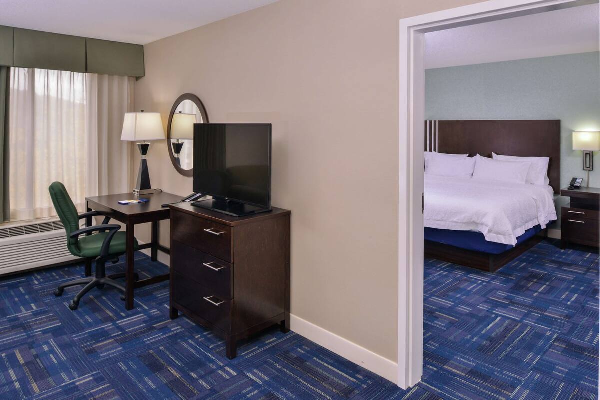 A hotel room with a desk, chair, TV, and a bed with white linens.