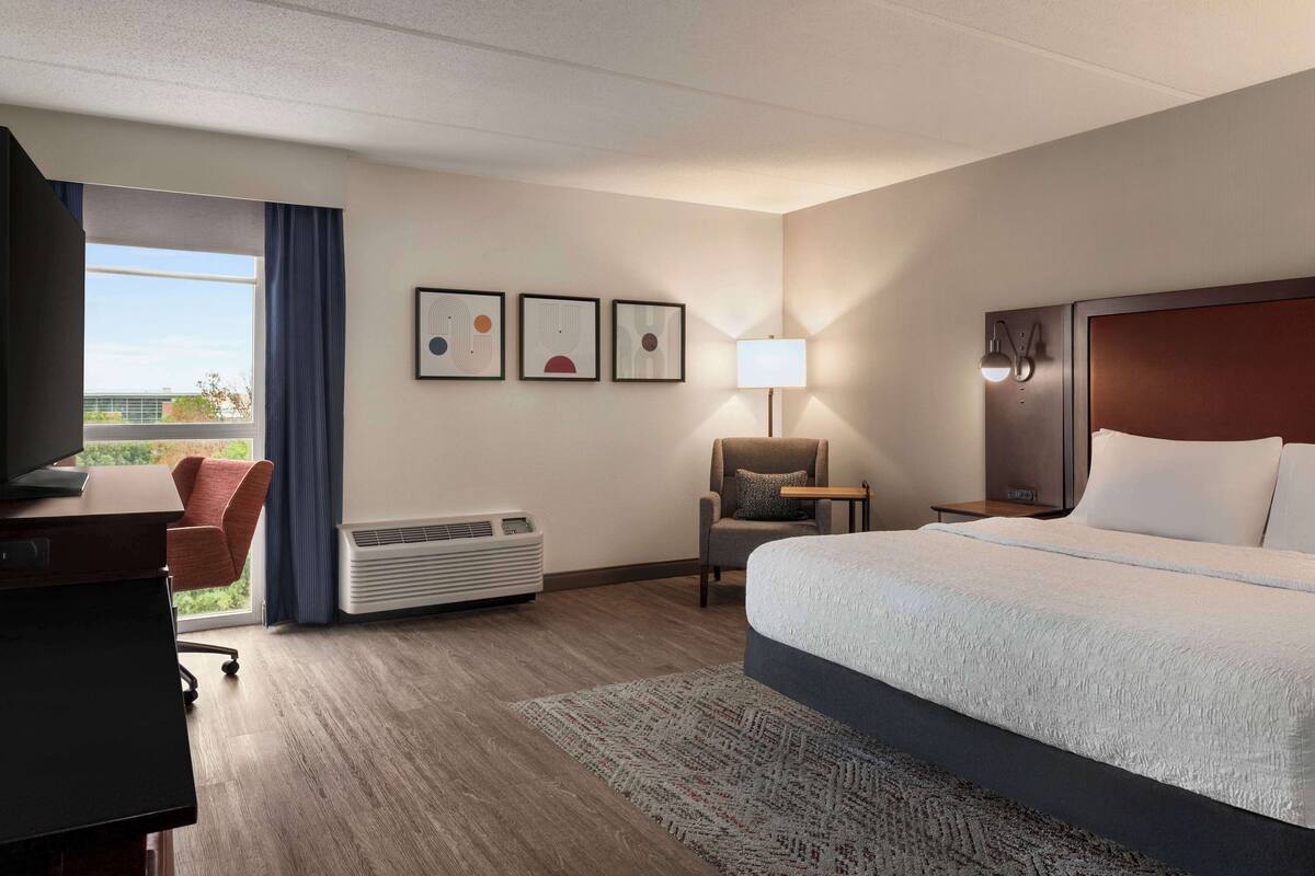 A hotel room with a bed, desk, chair, air conditioner, and framed artwork on the wall.