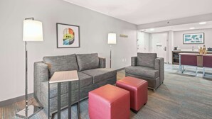 Ein modernes Hotelzimmer mit einem grauen Sofa, einem roten Pouf und einem kleinen Tisch. Zwei Stehlampen und ein gerahmtes Bild sind an der Wand zu sehen.