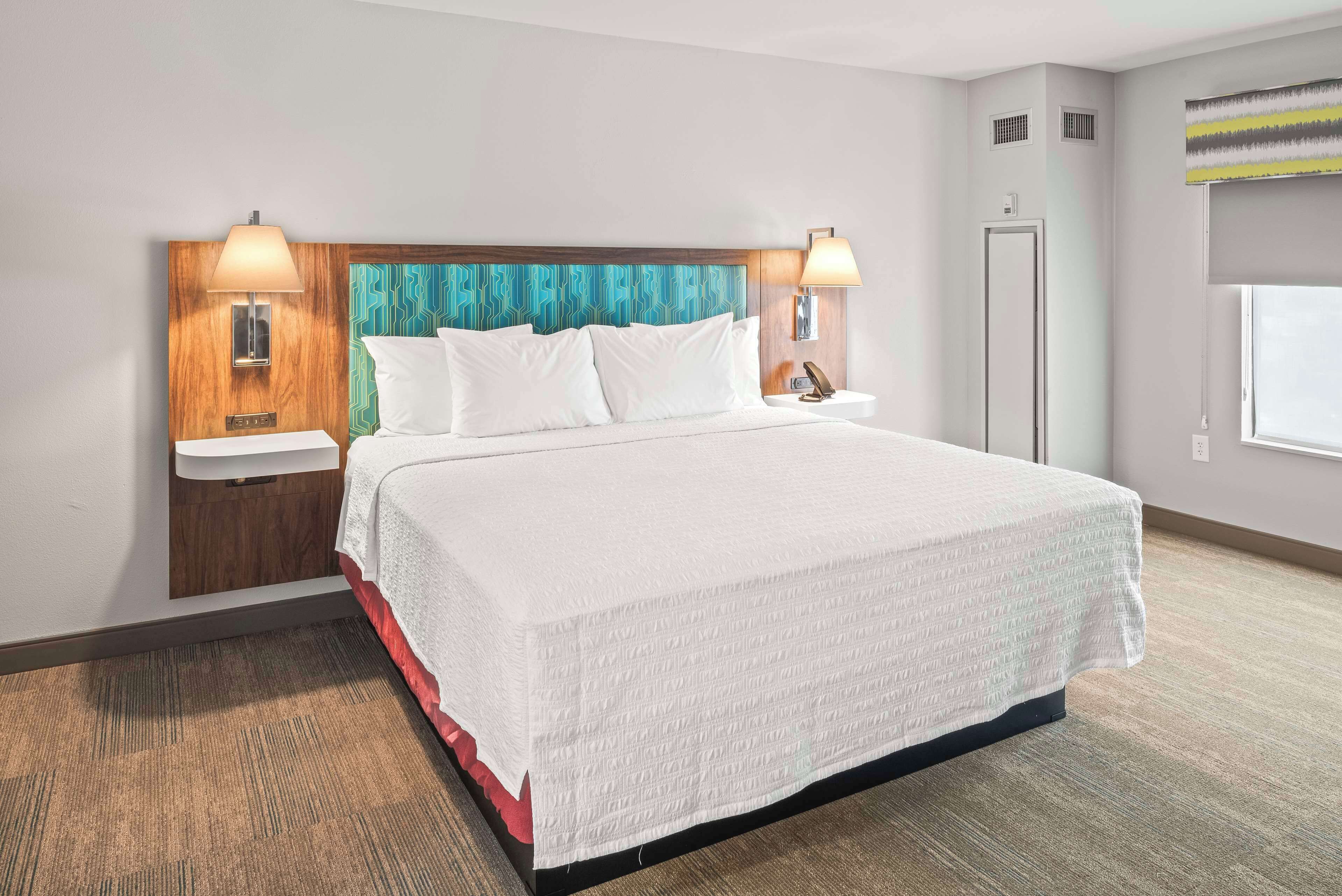 Une chambre d’hôtel avec un grand lit, deux tables de chevet avec des lampes, une tête de lit ornée d’un motif bleu, une fenêtre donnant sur un paysage agréable et une porte.