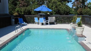 Una piscina rectangular con borde de ladrillo, rodeada por una terraza con baldosas blancas. Se ven sillas azules y un paraguas azul en el fondo.