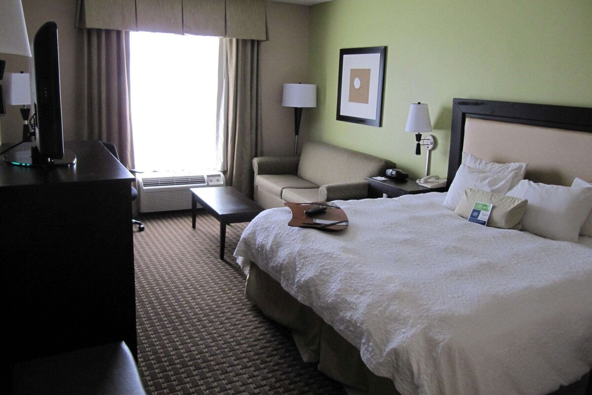 Habitación de hotel con cama, escritorio, silla, lámpara, un cuadro en la pared y ventana con cortinas.