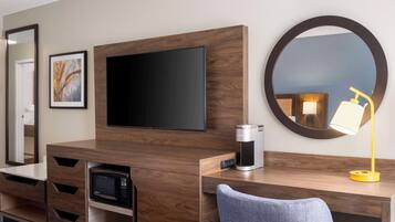 A hotel room with a wooden desk, a flat-screen TV, a coffee maker, a microwave, a mirror, and a lamp.