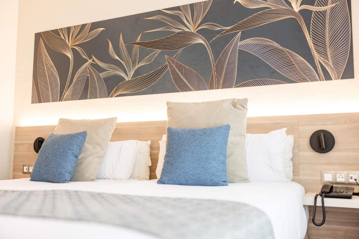 Habitación de hotel con cama, almohadas, teléfono en la pared y una obra de arte de gran tamaño con motivos de hojas sobre la cama.