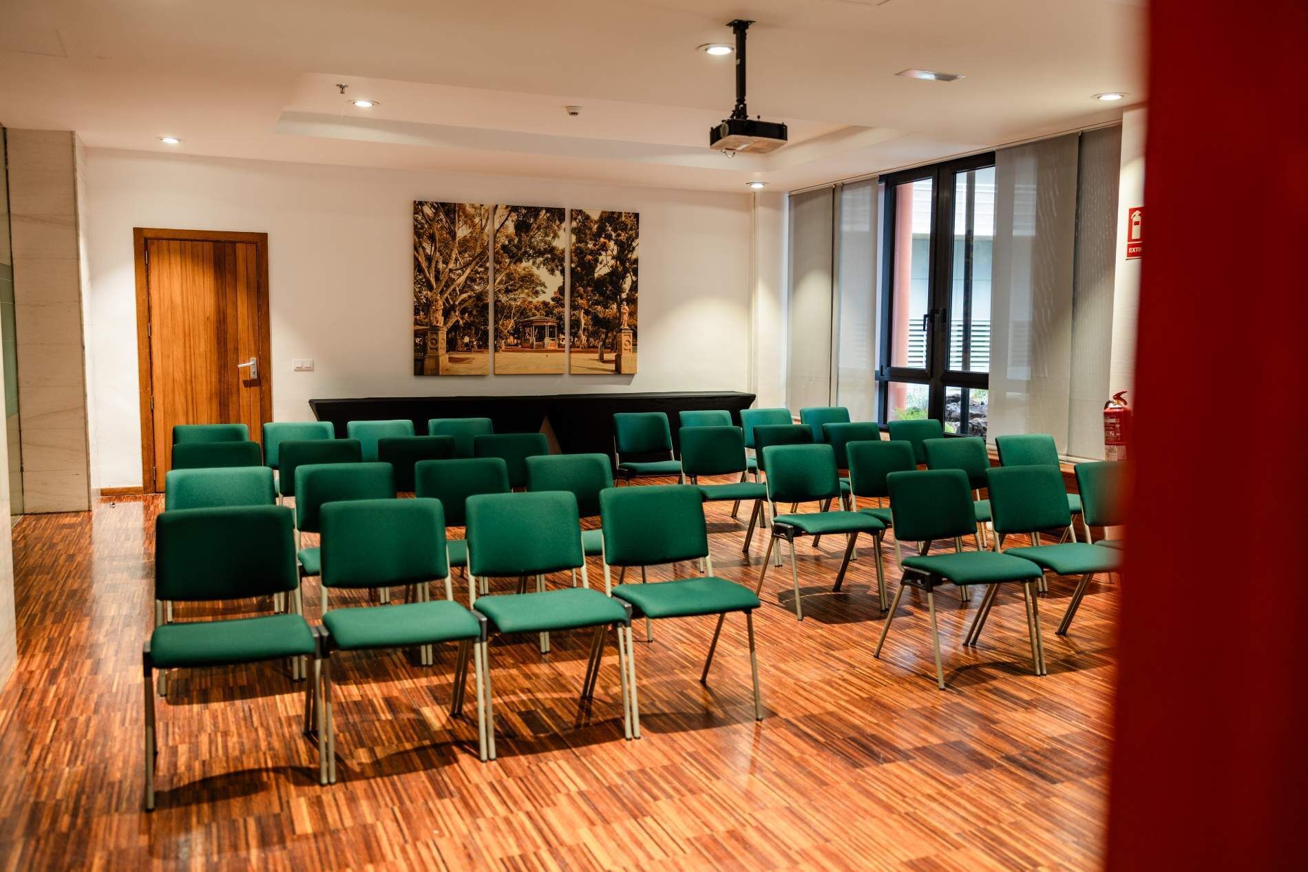 Una sala de conferències amb cadires verdes disposades en files, un paviment de fusta, un quadre gran a la paret i una porta.
