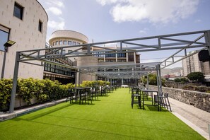 Un espacio de estar al aire libre con una estructura de pérgola metálica, césped artificial verde y vista a un edificio moderno con múltiples ventanales.