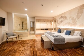 A modern hotel room with a wooden floor, a large bed with multiple pillows, a bedside table, a chair, a walk-in closet, and a bathroom with a bathtub and shower. - Hotel Escuela Santa Cruz (Santa Cruz de Tenerife)