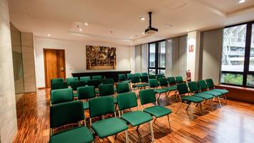 Uma sala de conferências com fileiras de cadeiras verdes, piso de madeira, uma grande janela e um projetor no teto.