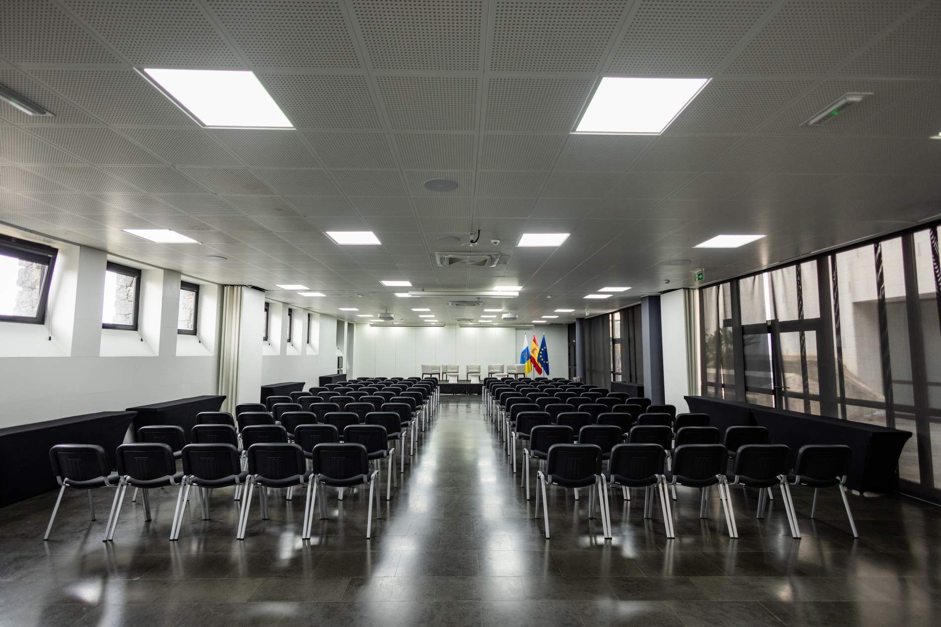 Un salón de conferencias con filas de sillas, un escenario y banderas.