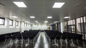 Un salón de conferencias con filas de sillas, un escenario y banderas.