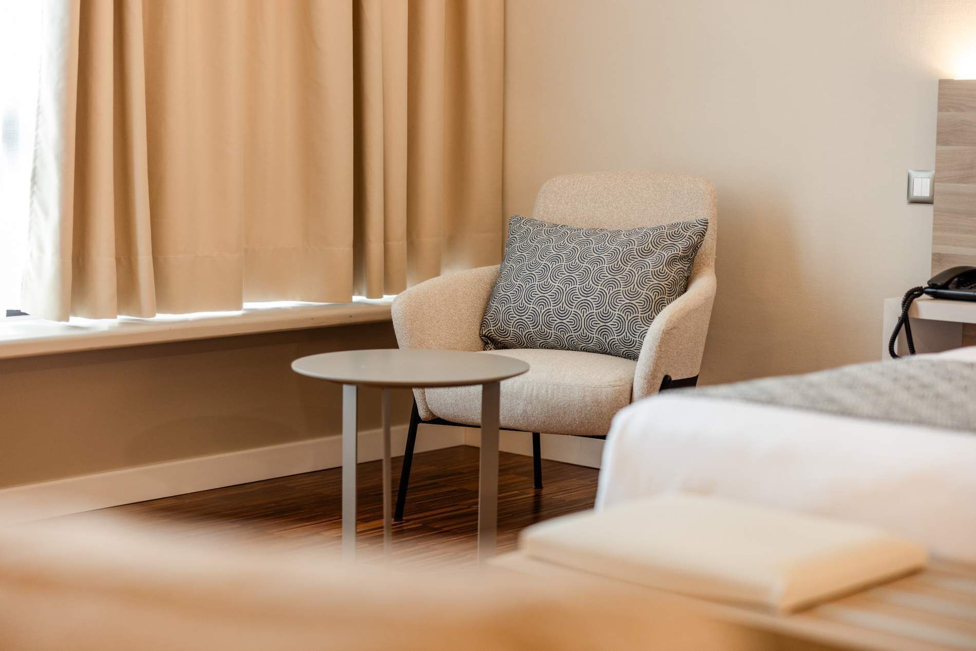 Een hotelkamer met een fauteuil, een klein rond tafeltje, een bed en een nachtkastje met een lamp.
