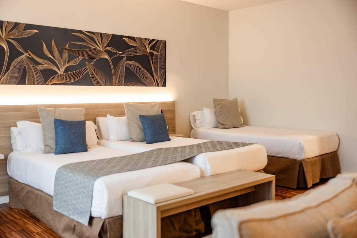 Habitación de hotel con dos camas, un banco, una obra de arte en la pared y suelo de madera.