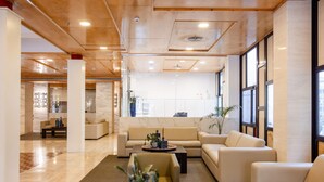 Un hall d’entrée spacieux au design moderne, comprenant un espace de détente central avec des canapés et une table basse, un comptoir d’accueil et de grandes fenêtres laissant entrer la lumière naturelle.