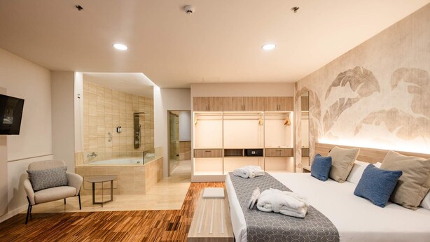 Une chambre d’hôtel moderne dotée d’un grand lit, d’un coin salon avec un fauteuil, d’une petite table, d’un dressing et d’une salle de bain avec douche et porte en verre.