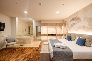 Une chambre d’hôtel moderne dotée d’un grand lit, d’un coin salon avec un fauteuil, d’une petite table, d’un dressing et d’une salle de bain avec douche et porte en verre.