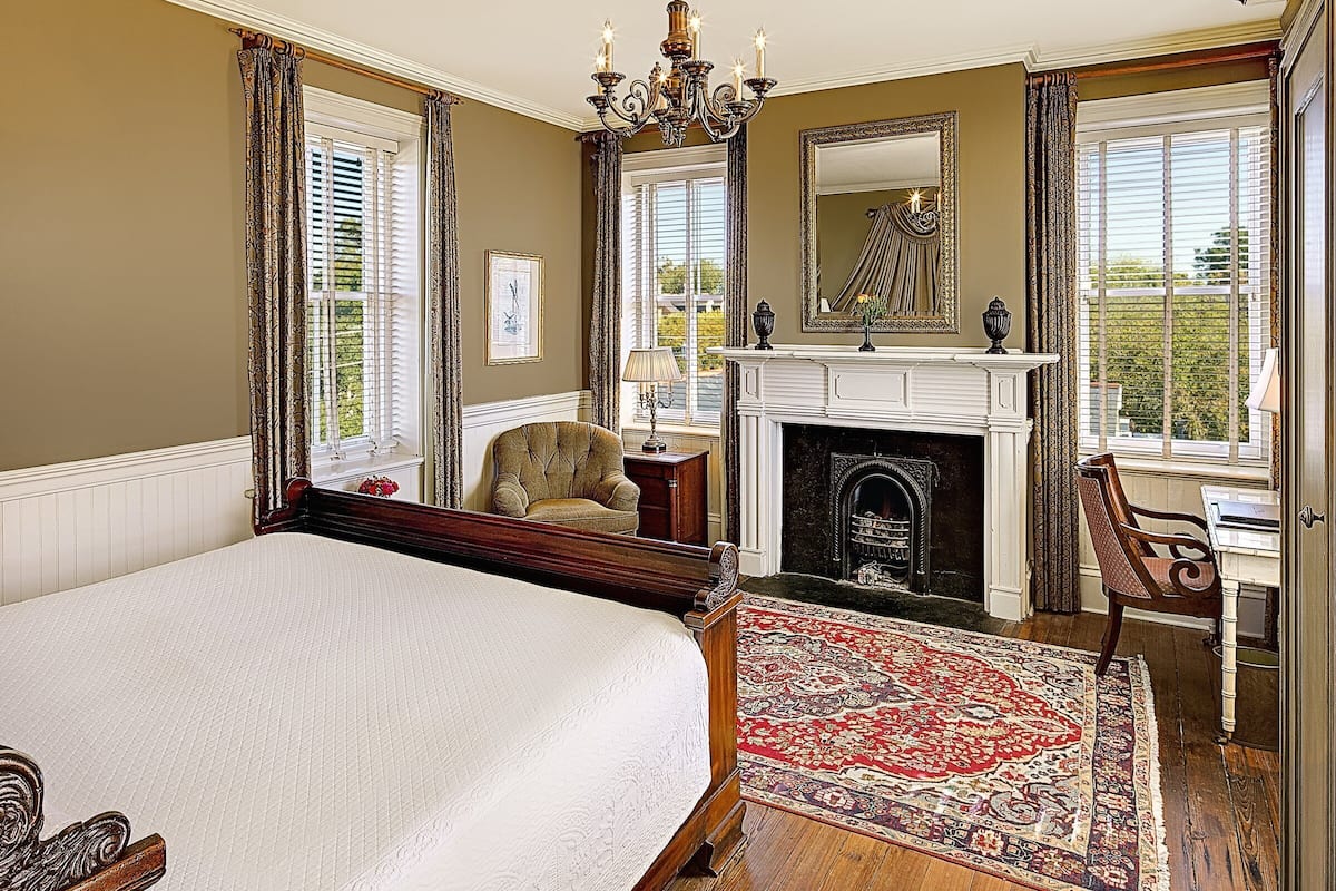A bedroom with a large bed, a fireplace, a chair, and a desk with a lamp.
