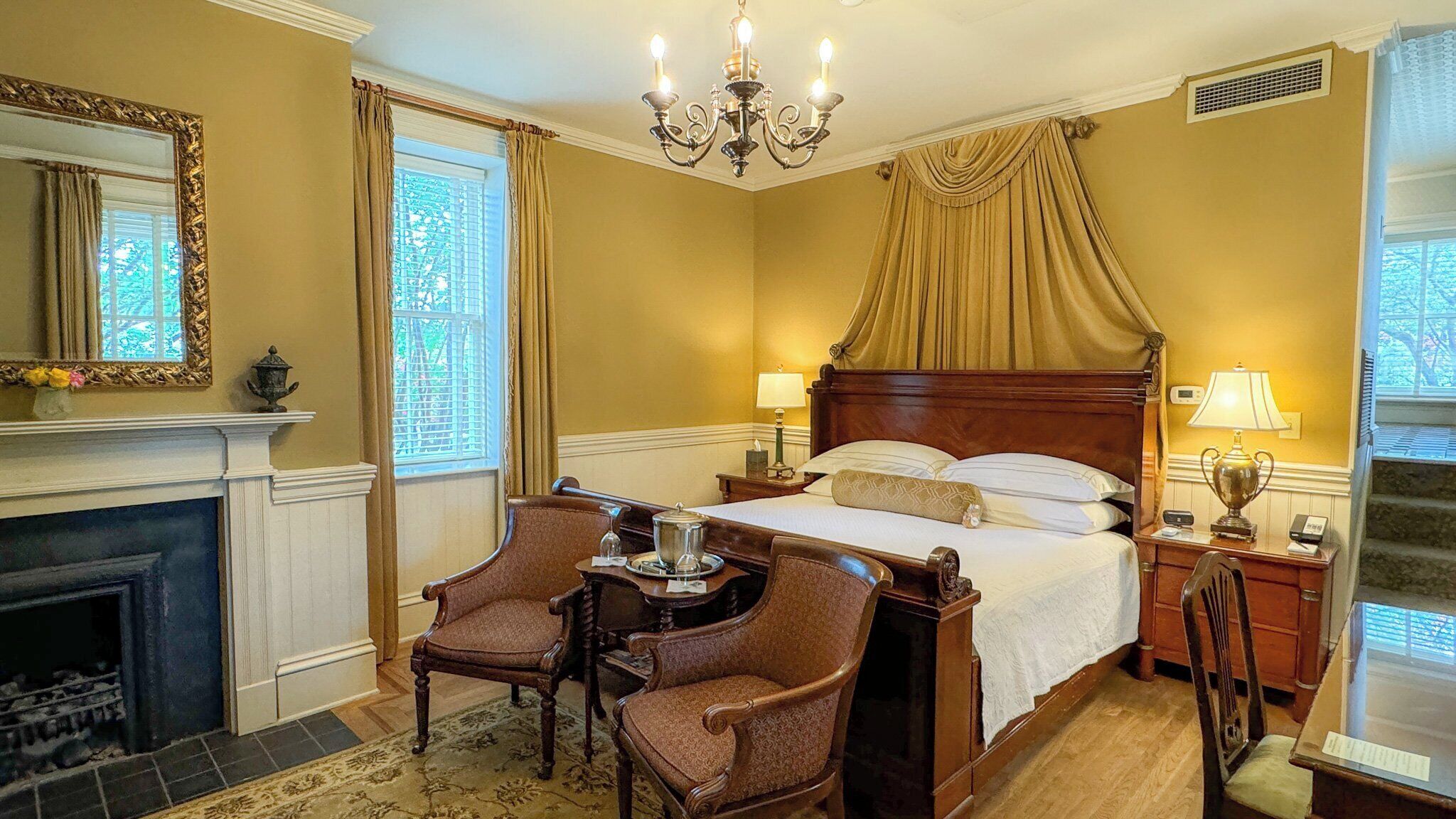 A classic hotel room with a large bed, two bedside tables, a chandelier, a fireplace, and a mirror.