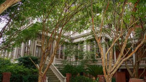 A courtyard with a staircase leading to a balcony, surrounded by lush greenery and a brick wall.