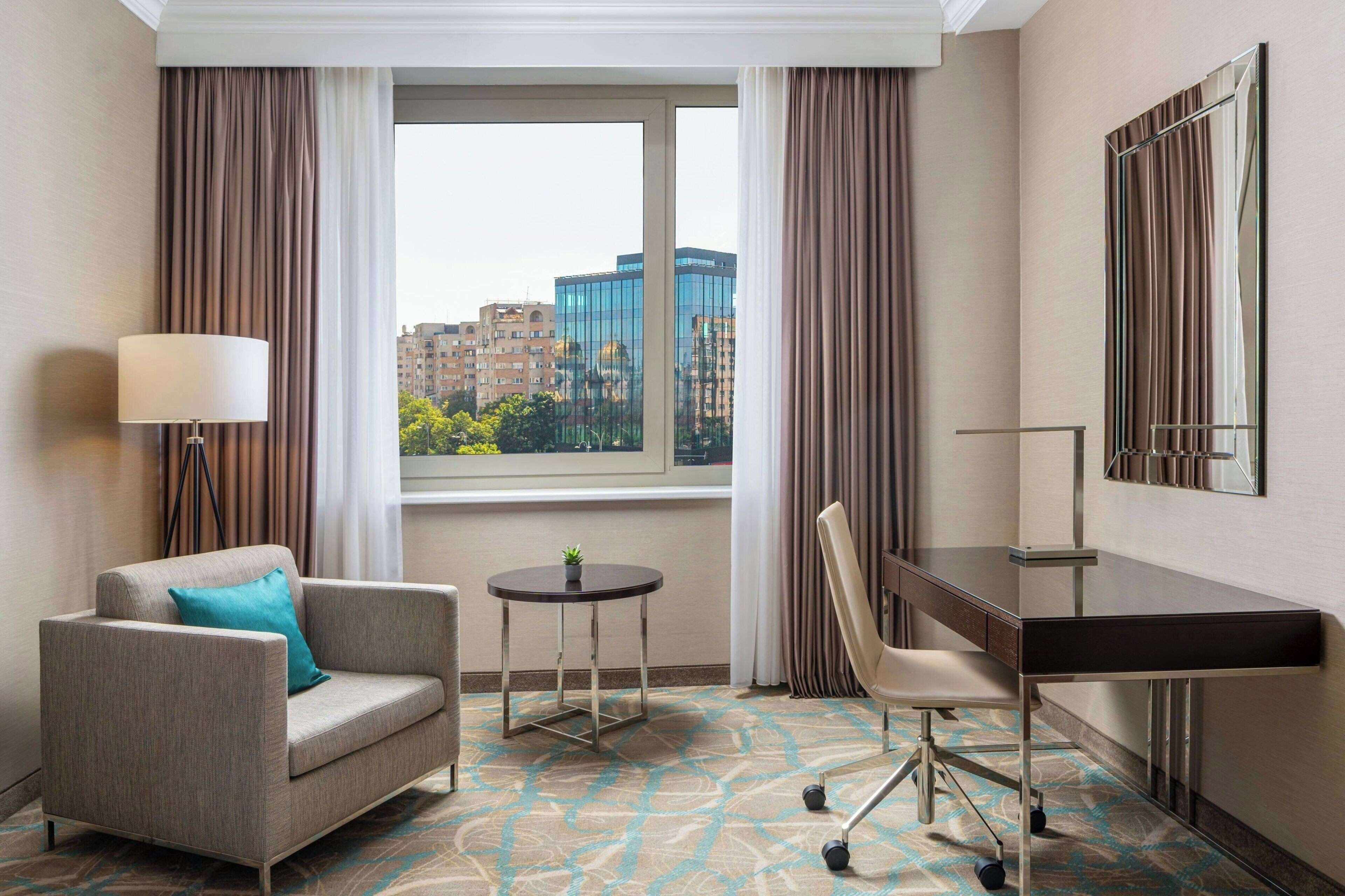 A hotel room with a sofa, a desk, a chair, a lamp, a small table with a plant, and a large window with a city view.