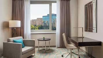 A hotel room with a sofa, a desk, a chair, a lamp, a small table with a plant, and a large window with a city view.