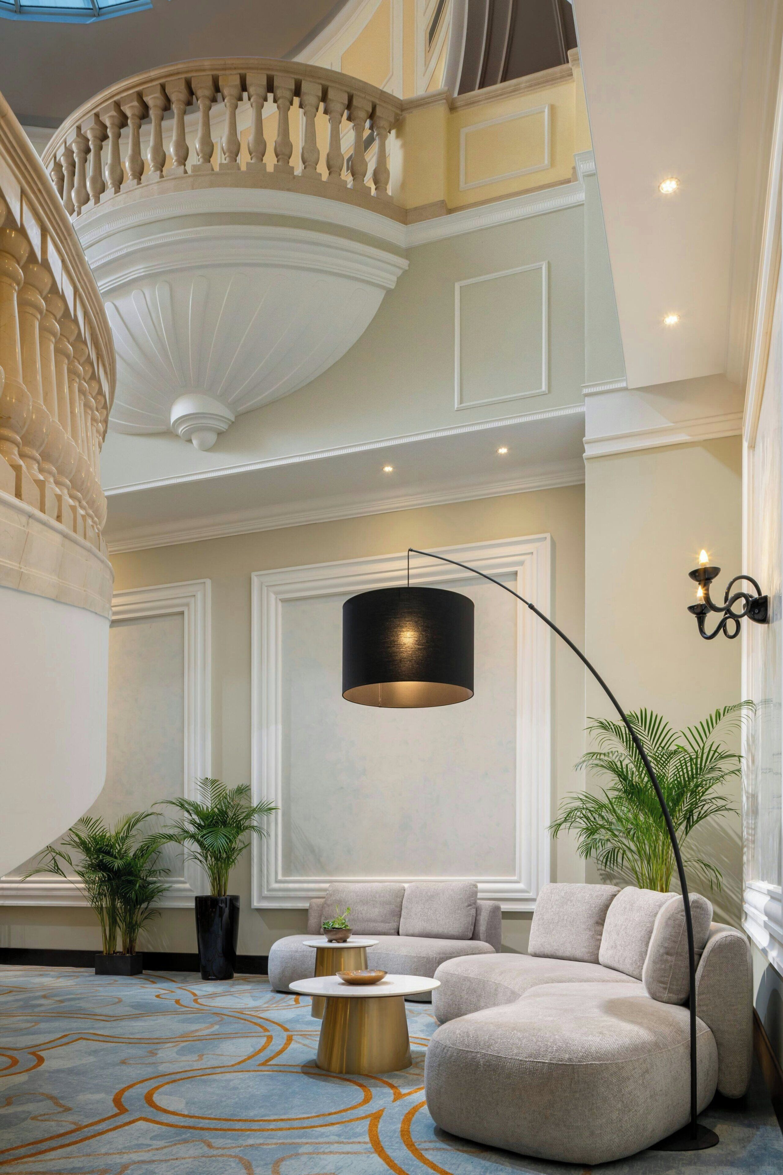A spacious lobby with a curved sofa, a round coffee table, and a large floor lamp. There are potted plants and a staircase with a balustrade.