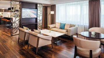 A modern hotel room with a sofa, chairs, a dining table, a TV, and a large window with curtains.