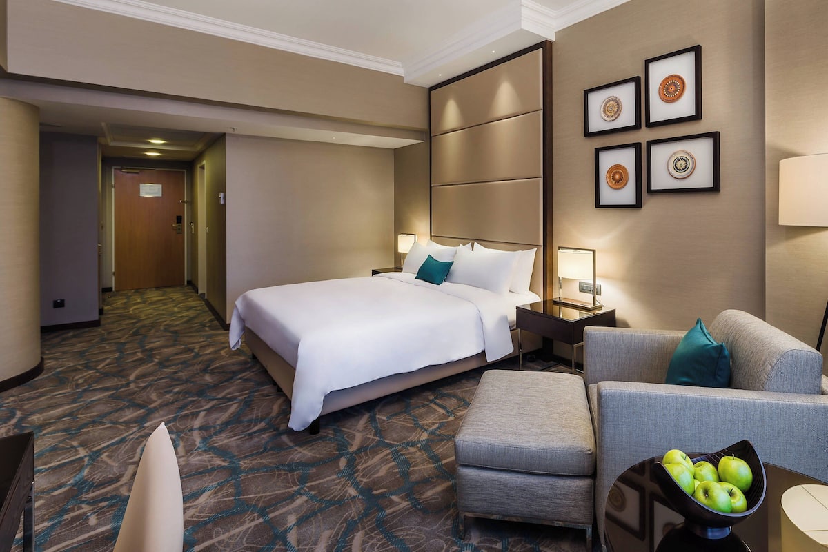 A hotel room with a large bed, a sofa, a small table with a bowl of fruit, and a wall with framed pictures.