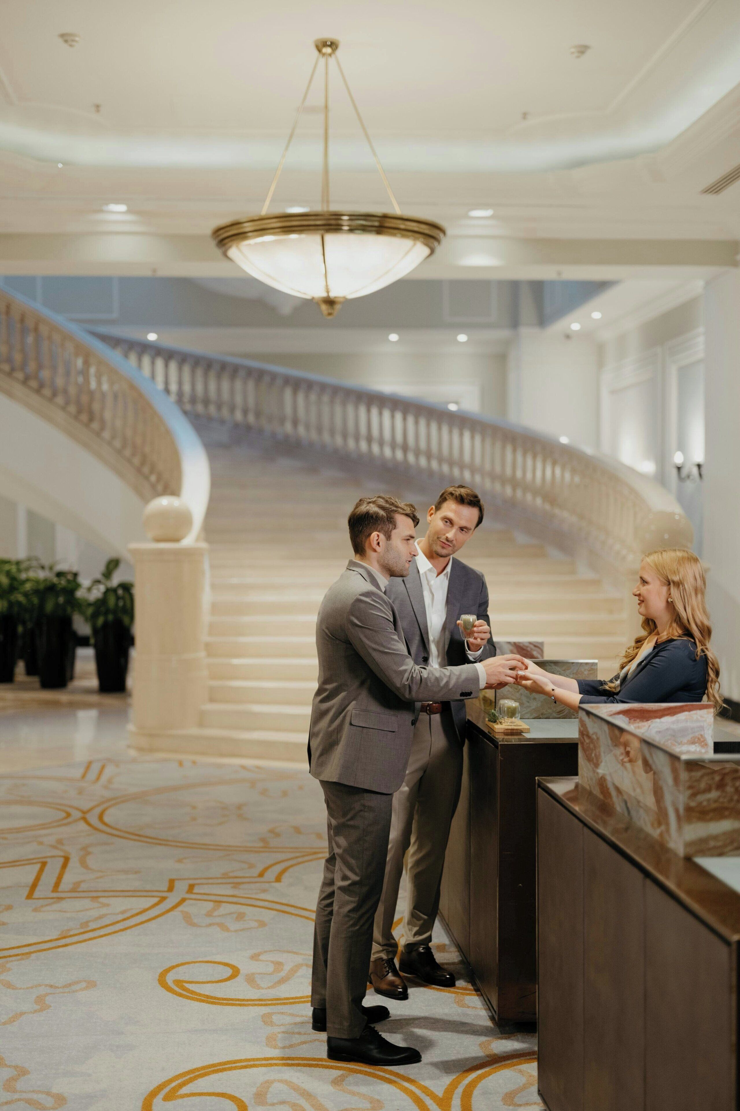 A reception area with a marble floor, a grand staircase in the background, and a chandelier.