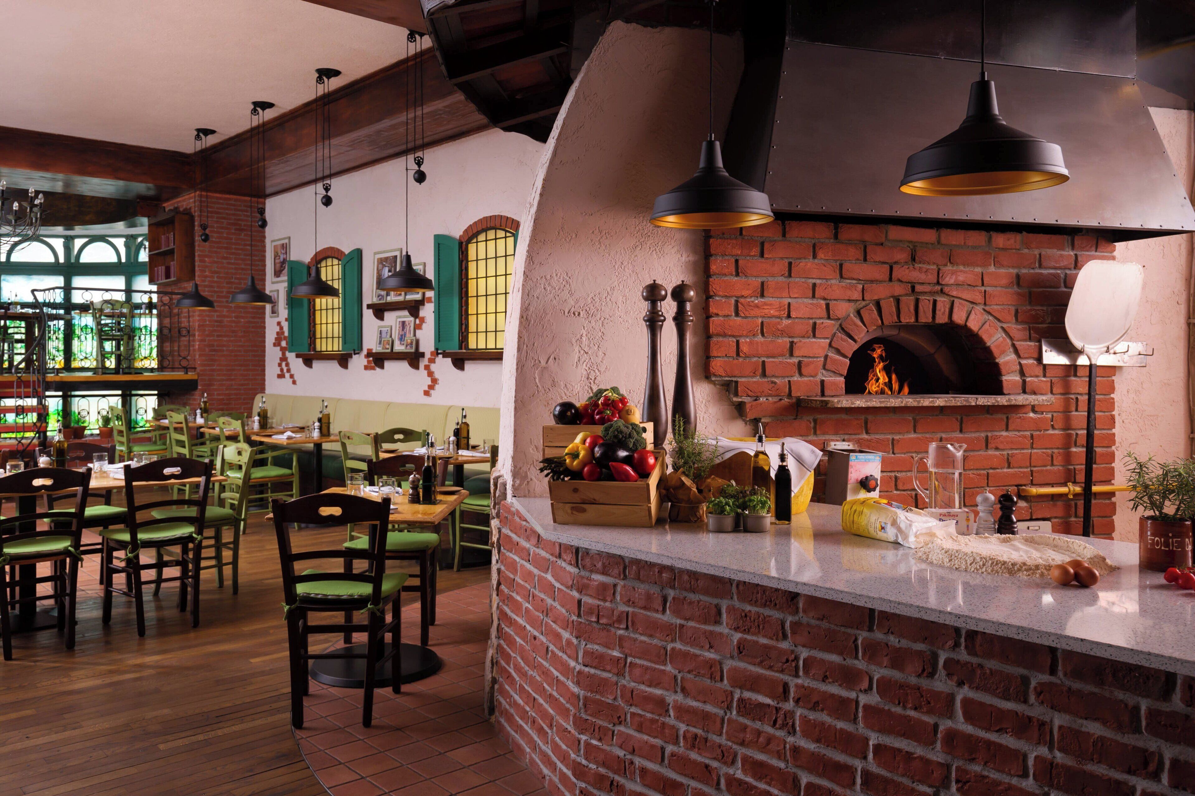 L’intérieur d’un restaurant avec une cheminée en briques, des tables en bois, des chaises vertes et des lampes suspendues.