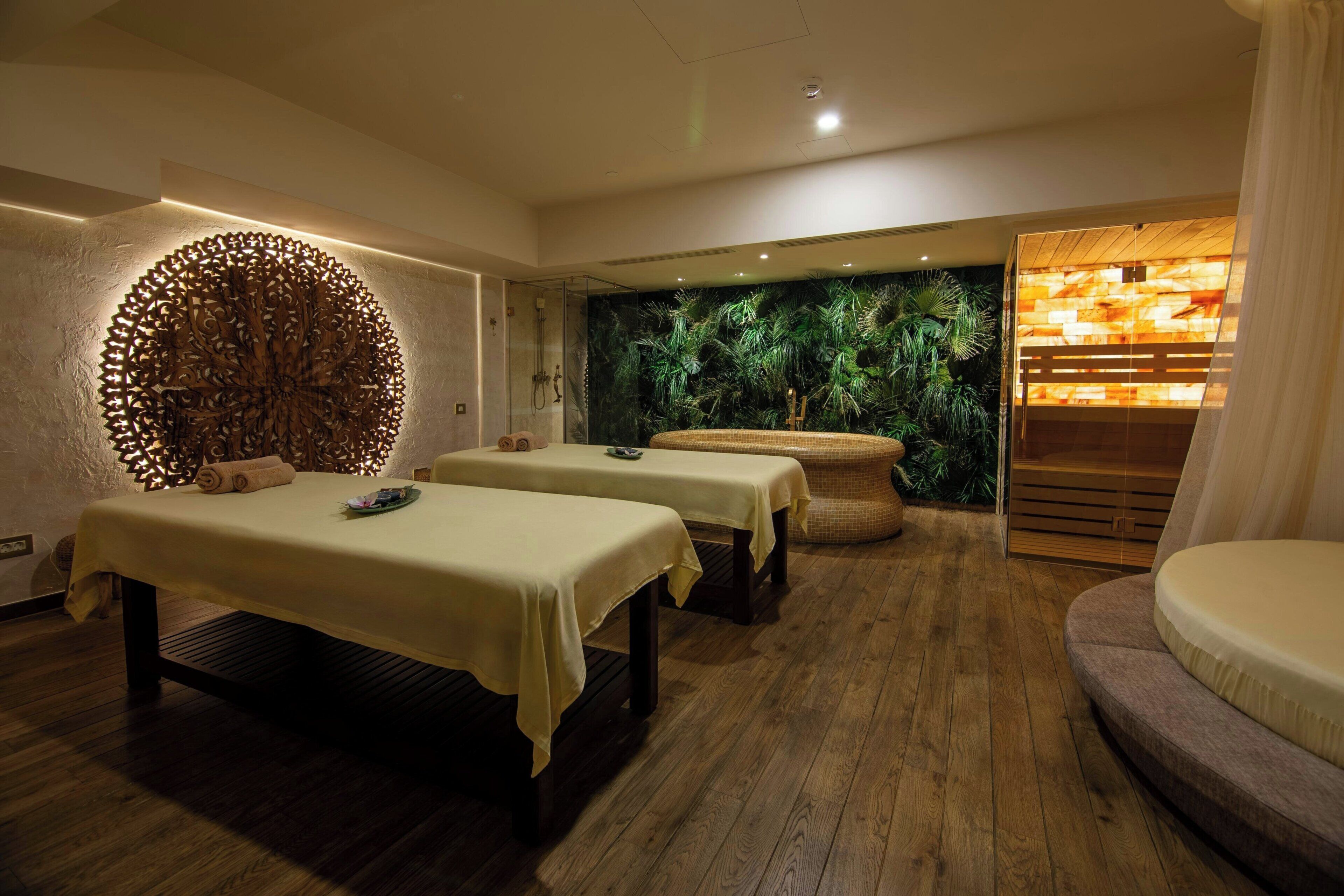 Uma sala de spa com duas mesas de massagem, uma parede com design circular em madeira, uma parede com jardim vertical e um banco de madeira.