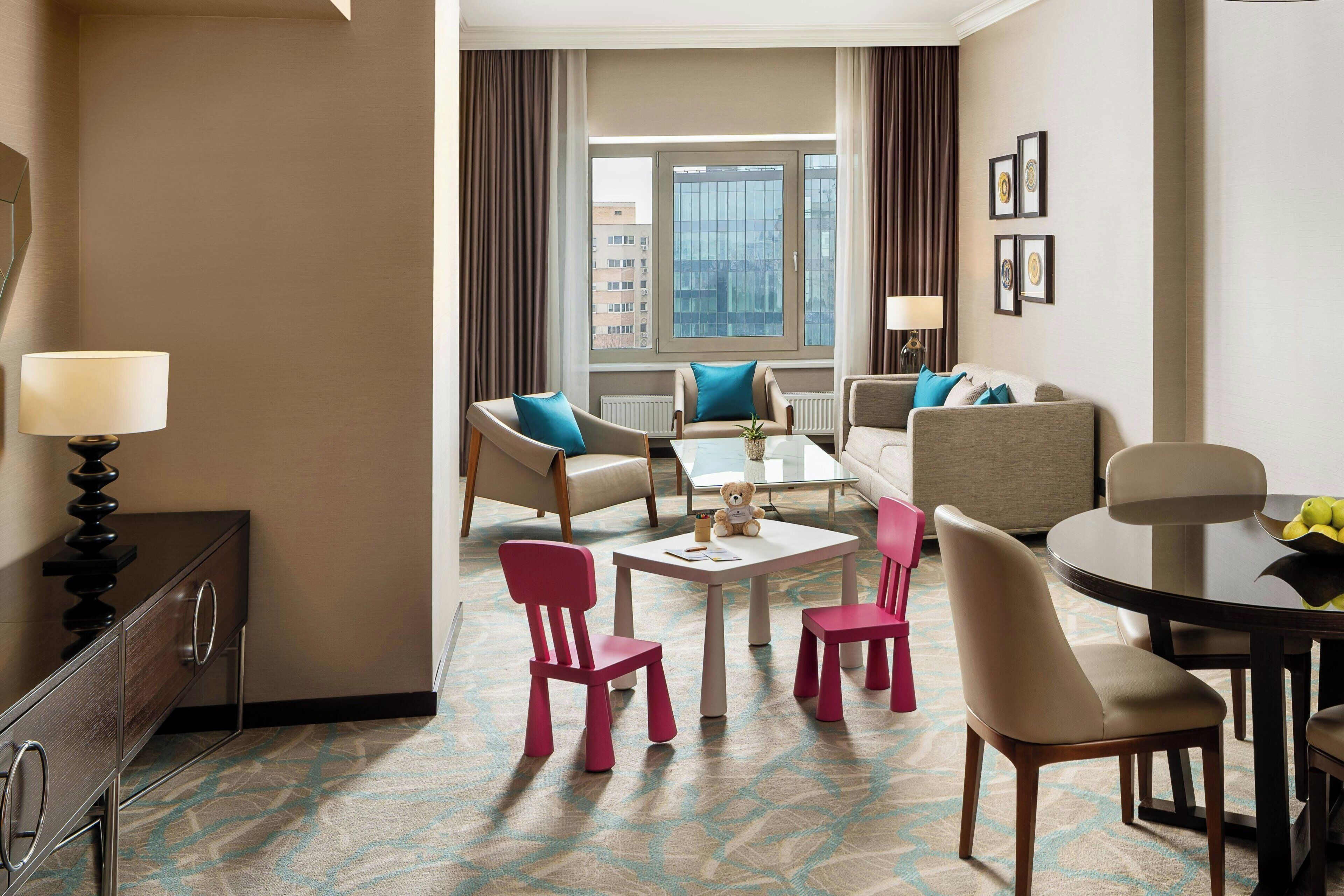 A hotel room with a seating area, a dining table, and a view of the cityscape.
