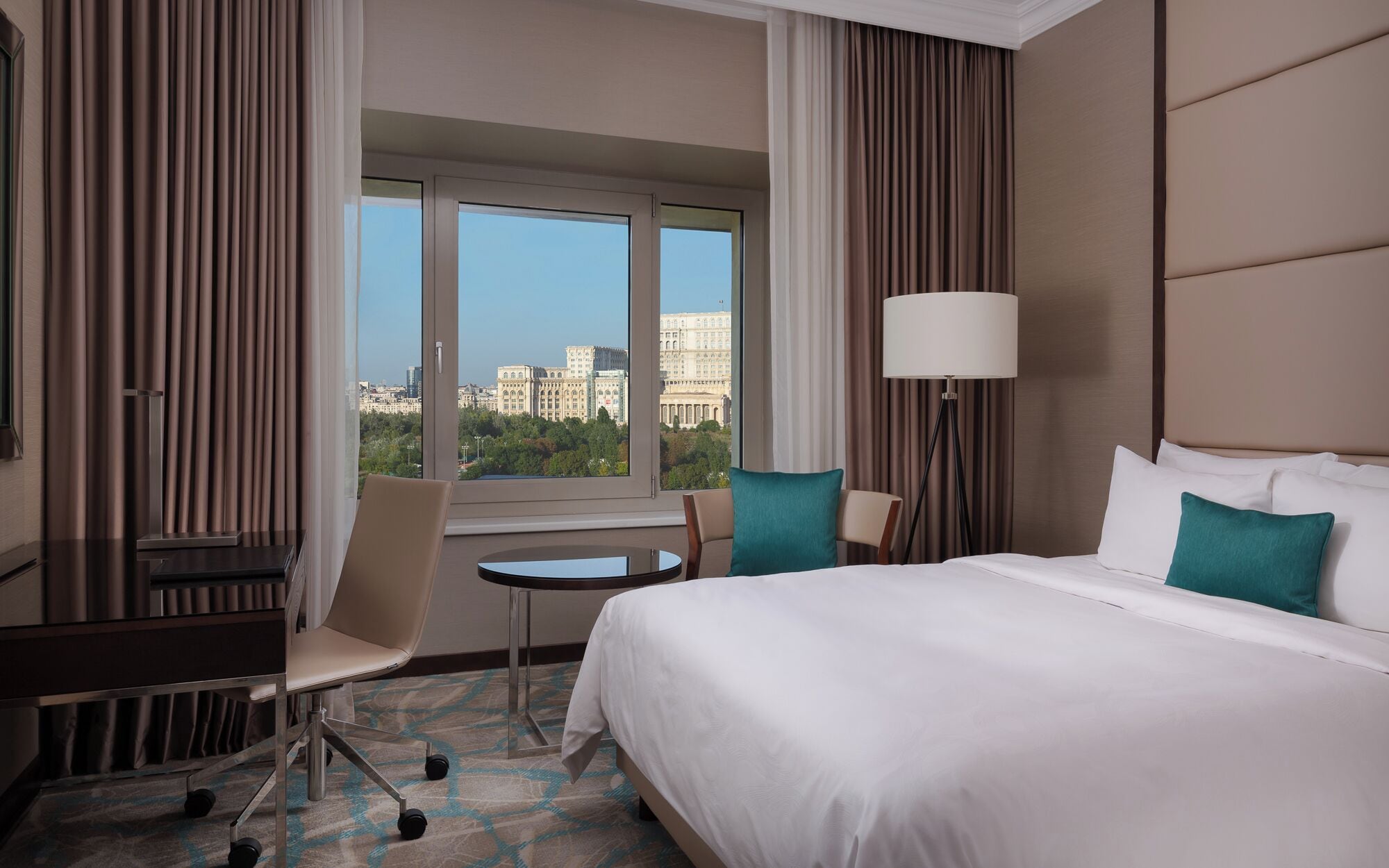 A hotel room with a bed, desk, chair, lamp, and a view of a cityscape through large windows.