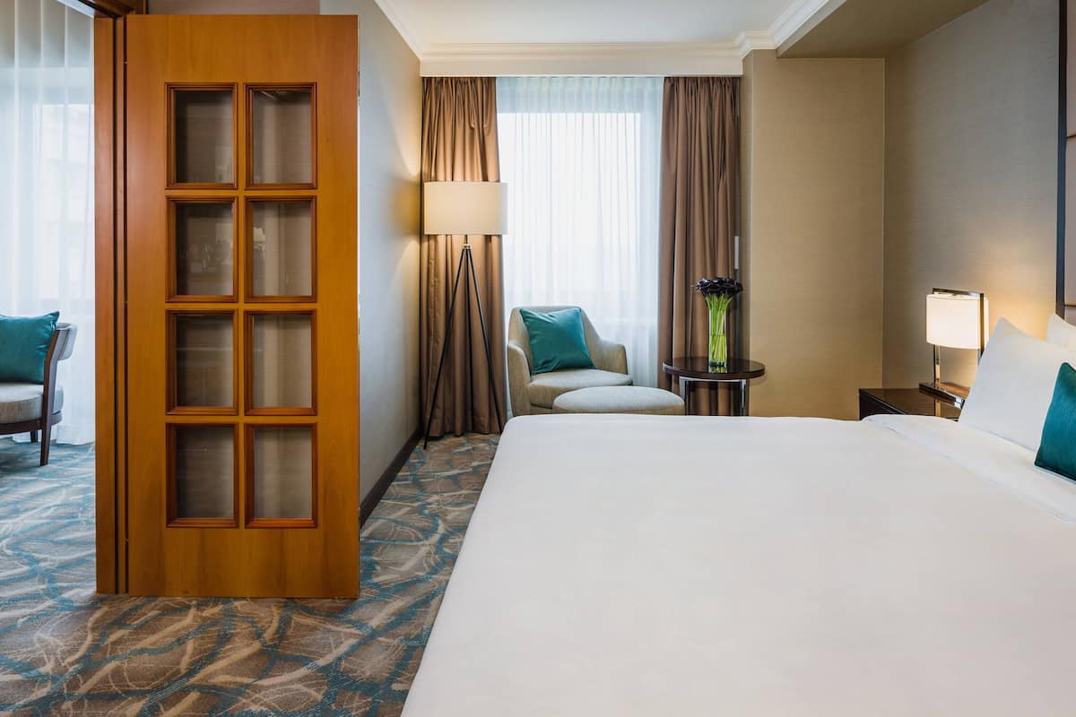 A hotel room with a large bed, a wooden door with glass panels, a chair, a small table with a lamp, and a nightstand with a vase.