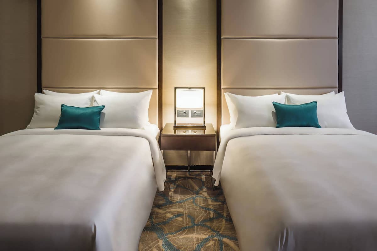 A hotel room with two beds, each with white linens and teal pillows, a nightstand with a lamp, and a patterned carpet.