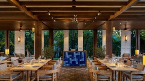 Een restaurant met een lange houten tafel en stoelen, een bar met blauwe tegels en grote ramen met uitzicht op bomen.