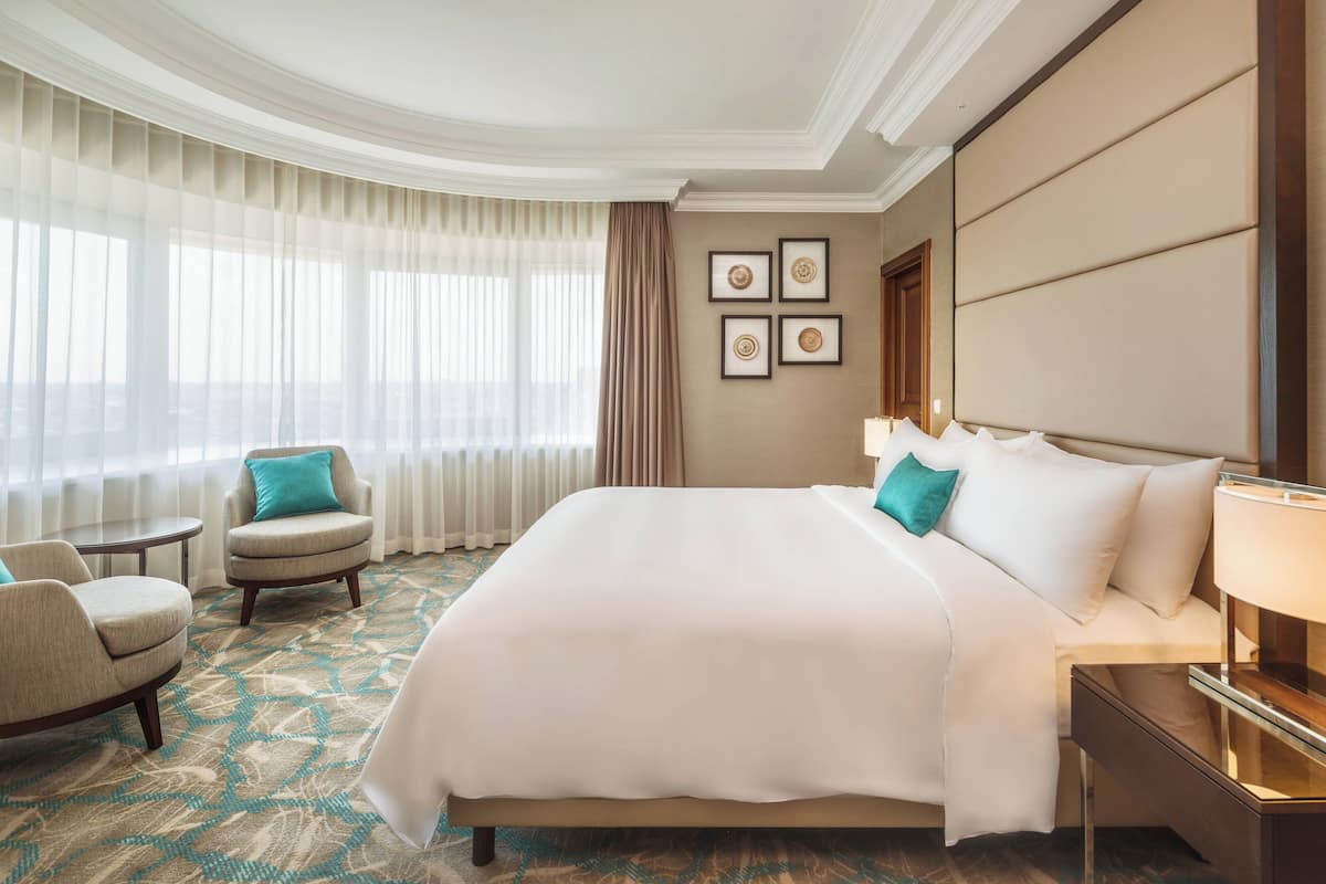 A hotel room with a large bed, two armchairs, a small table, and a nightstand with a lamp.