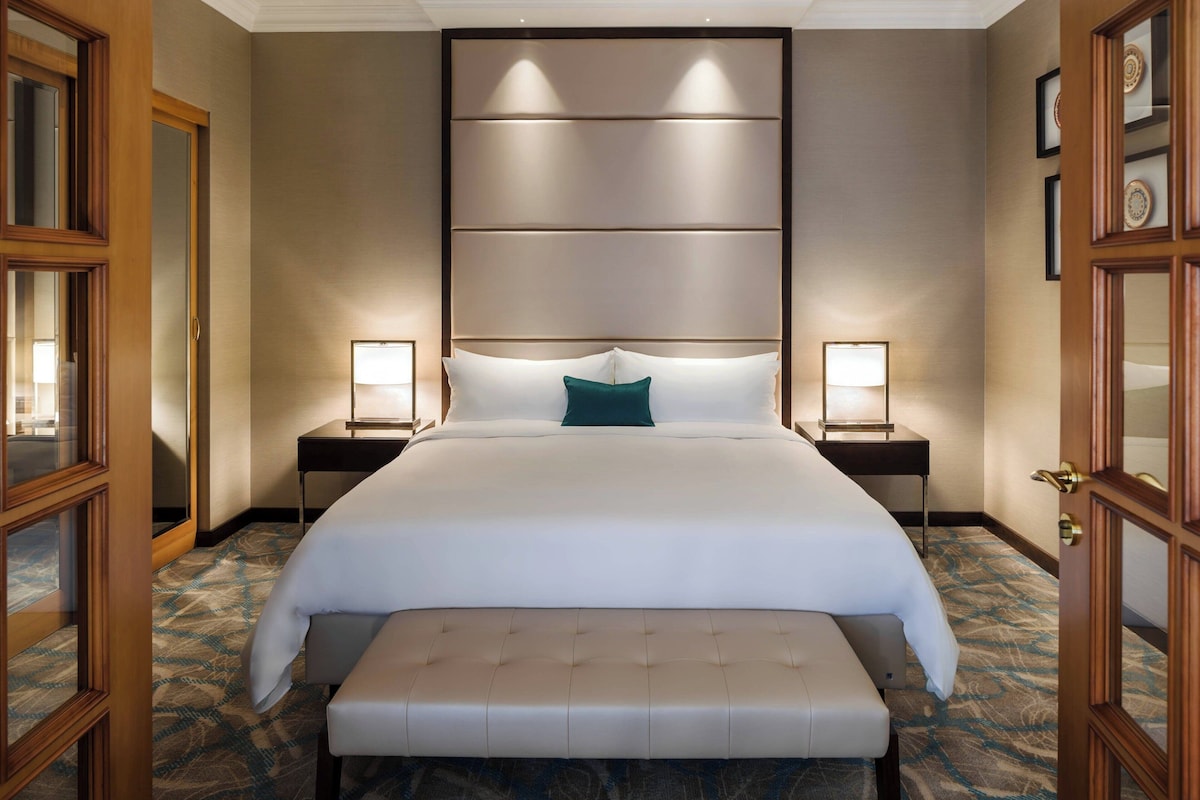 A hotel room with a large bed, two bedside tables with lamps, a bench at the foot of the bed, and a patterned carpet.