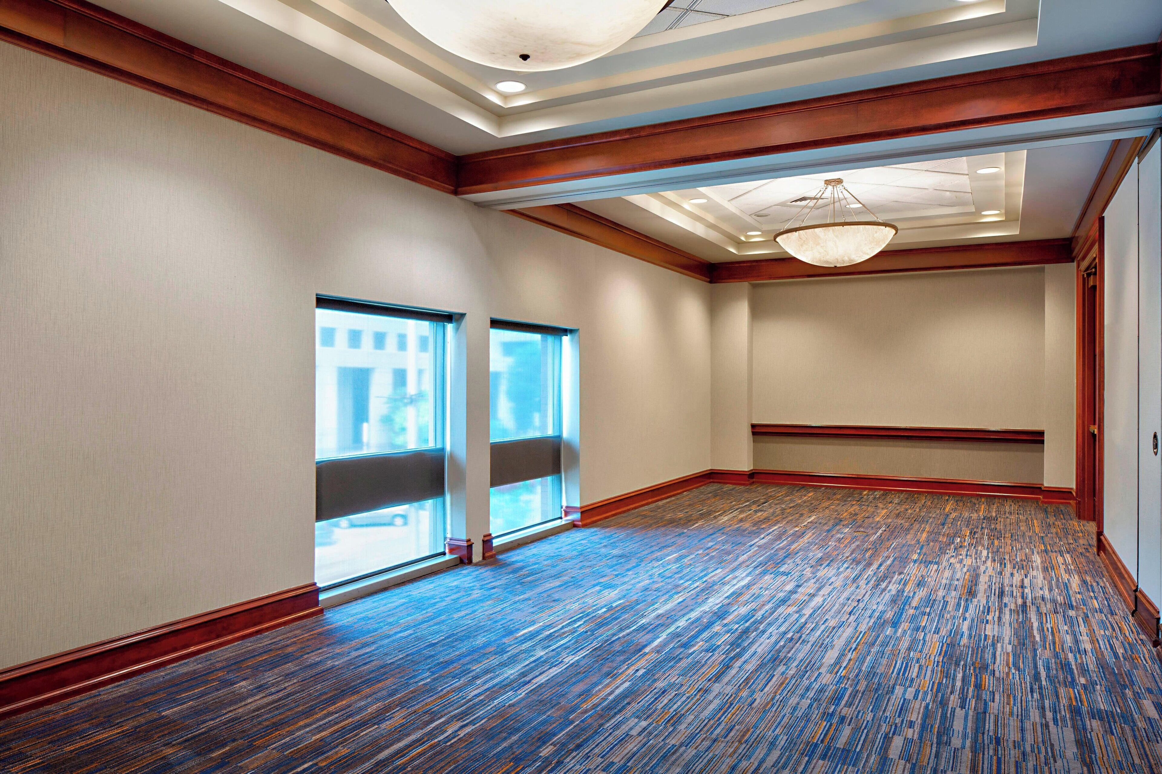 A large, empty room with a patterned carpet, wooden trim, and a ceiling with recessed lighting and a chandelier.