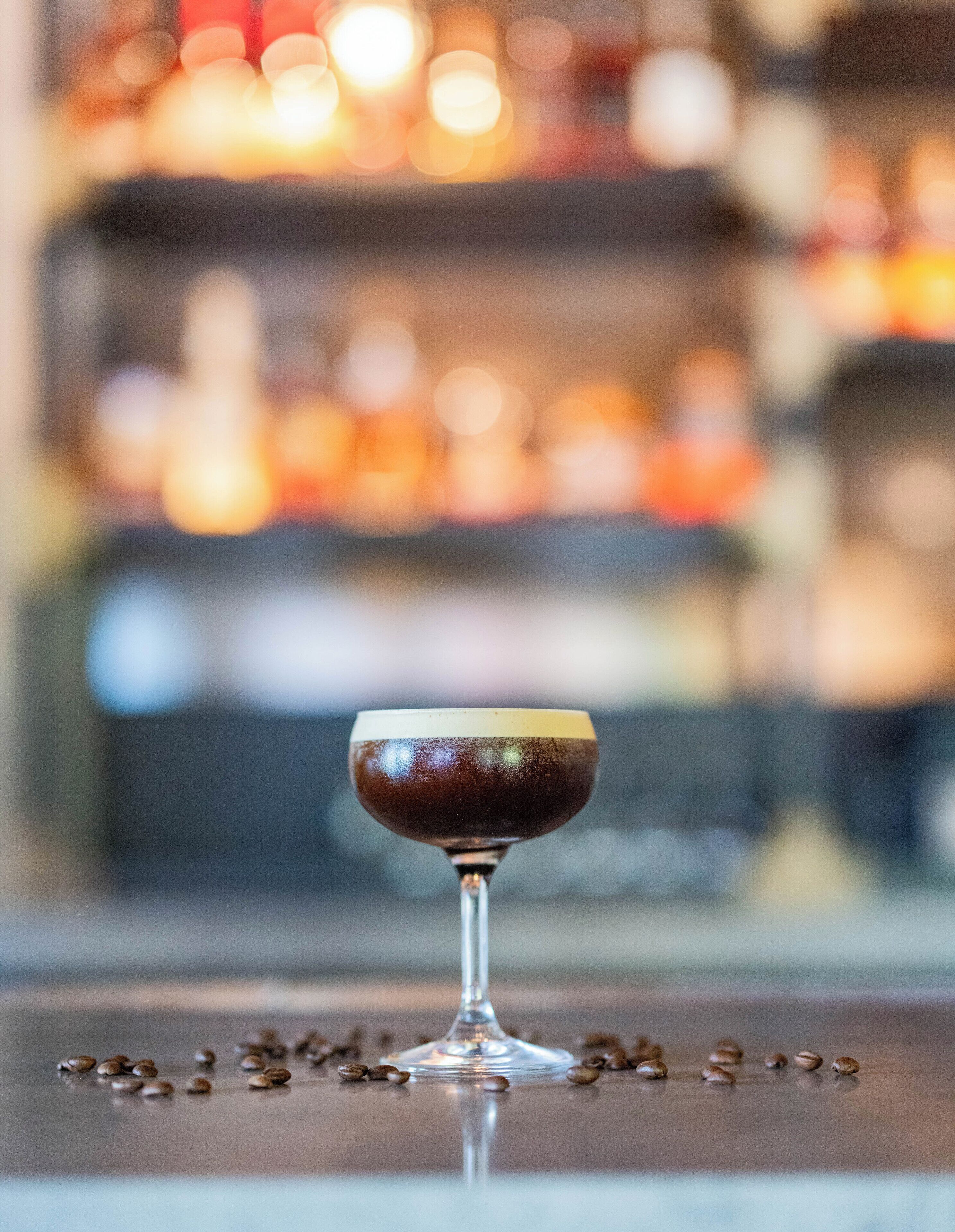 Ein Glas dunkler Flüssigkeit auf einer reflektierenden Oberfläche, umgeben von Kaffeebohnen.